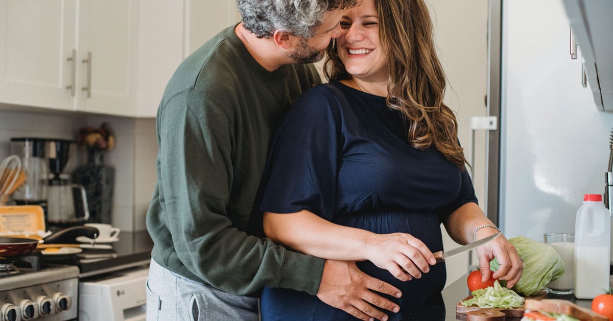 Middle-aged male wraps his hands around his pregnant wife's hips as she smiles back at him. They are in a kitchen and she is chopping lettuce and tomatoes on a wooden cutting board for a meal. | Optimizing Nutrition and the Prenatal Diet for a Healthy Pregnancy