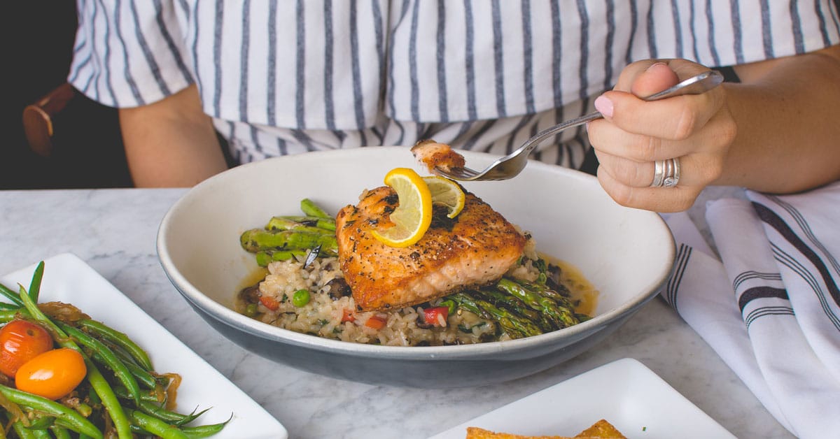 Women in striped blue and white dress sits at a table eating a large filet of salmon on top of rice and asparagus and garnished with lemon slices | Optimizing Nutrition and the Prenatal Diet for a Healthy Pregnancy with Omega 3s