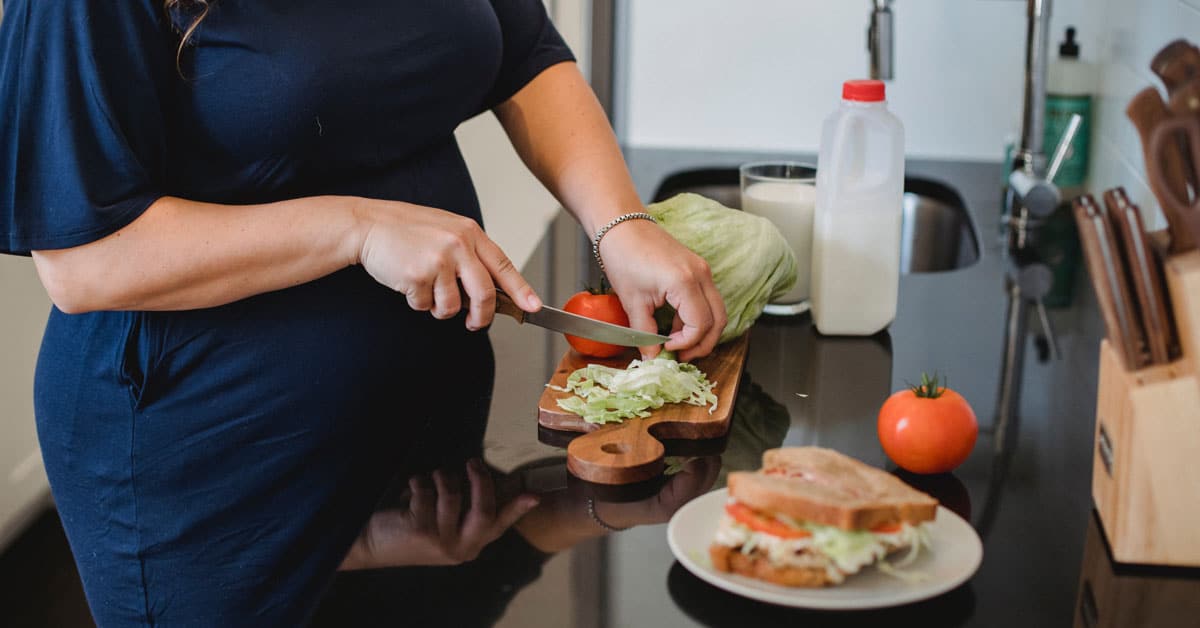 Pregnant woman in navy dress chops lettuce and tomato on a wooden cutting board with a sandwich in the foreground and glass of milk in the background | Optimizing Nutrition and the Prenatal Diet for a Healthy Pregnancy with Dairy